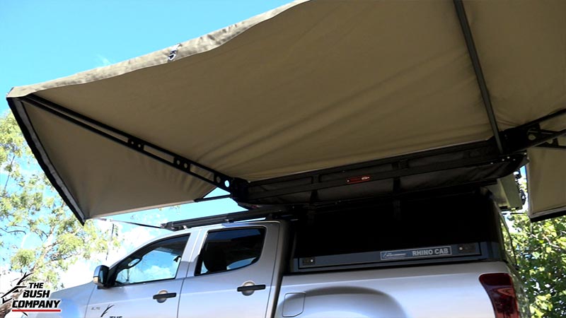 White pickup truck under tan rooftop tent and awning, trees and sky visible.