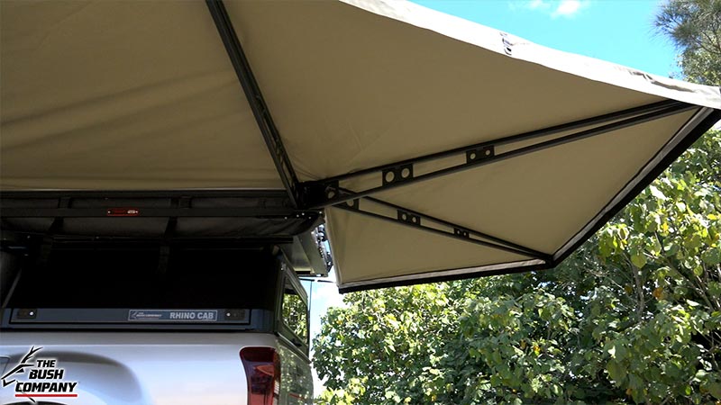 White pickup truck under tan rooftop tent and awning, trees and sky visible.