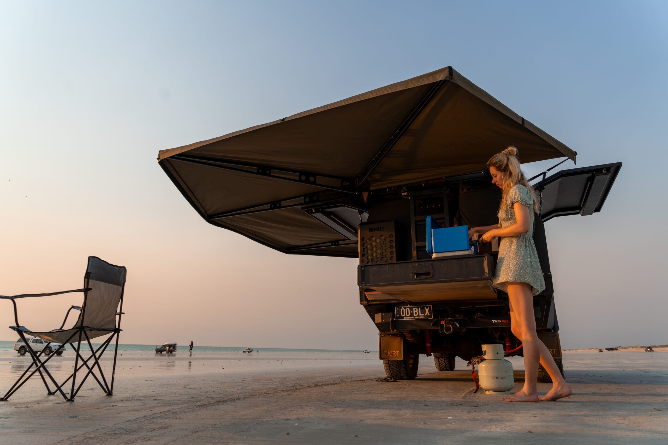 Woman at back of camper truck on beach, open awning, empty chair, ocean in distance.