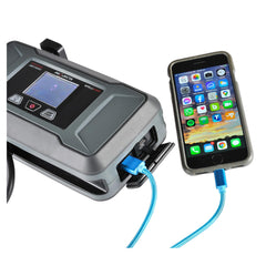 A portable jump starter unit charging a smartphone via a blue USB cable, with the phone screen displaying various app icons. The jump starter screen shows a battery icon, indicating it's functioning as a power bank.