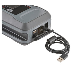 Close-up of a portable jump starter with a USB cable plugged into one of its ports, showing multiple output options including a standard USB and a 12V DC socket. The coiled USB cable is connected and ready for charging or power delivery.