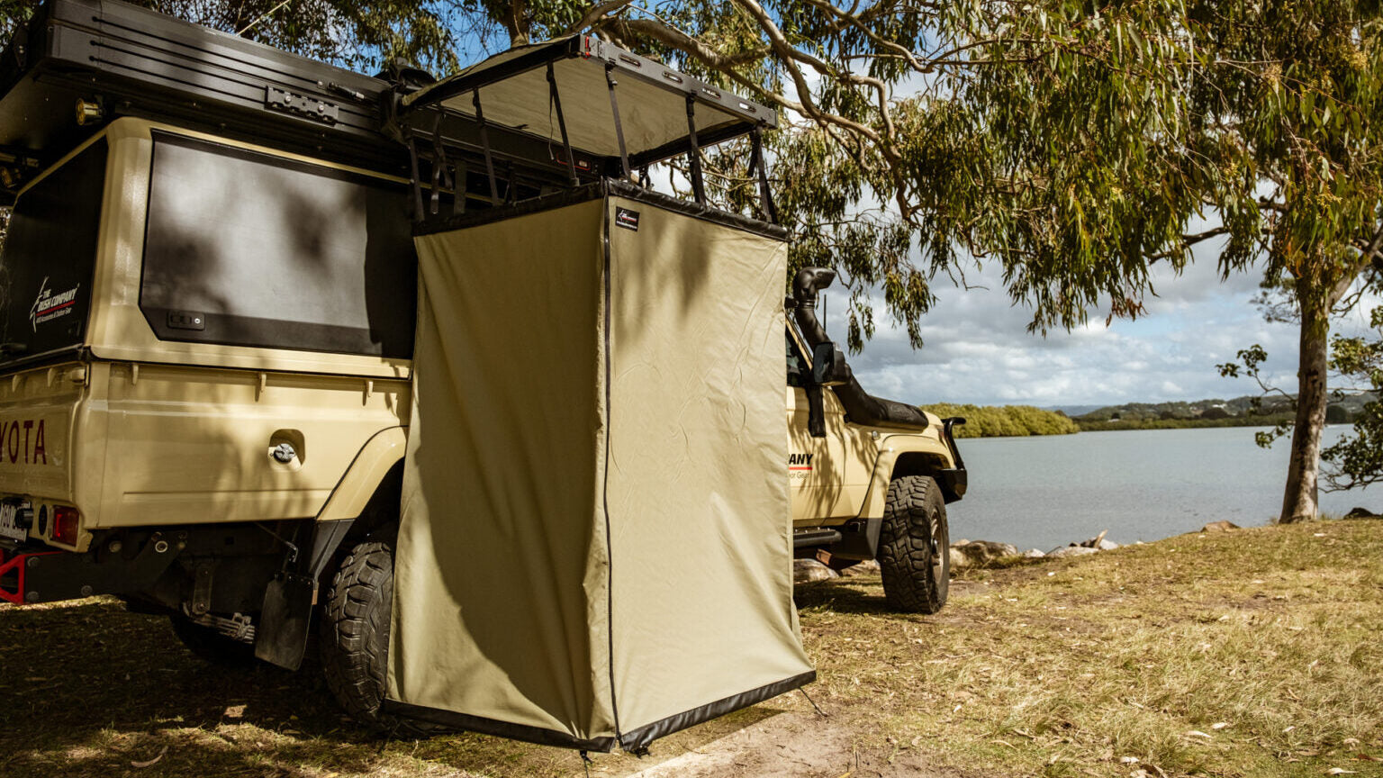 Beige off-road vehicle near lake, rooftop tent, side awning, trees, cloudy sky.