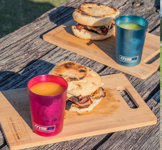 Outdoor breakfast setup with sandwiches and colorful MSA cups on wooden boards.