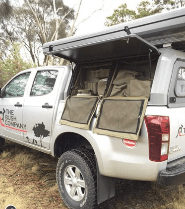 White pickup with rooftop tent and storage compartments, parked by trees.