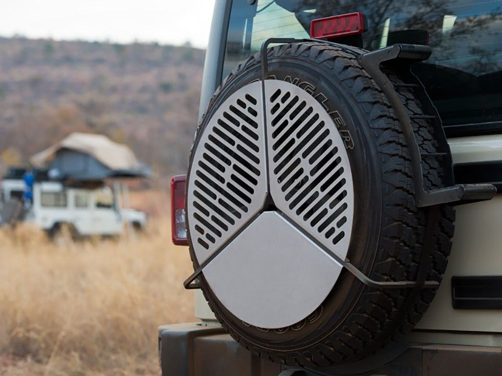 Metal shield on spare tire, vehicle with roof tent in background, grassy hills.